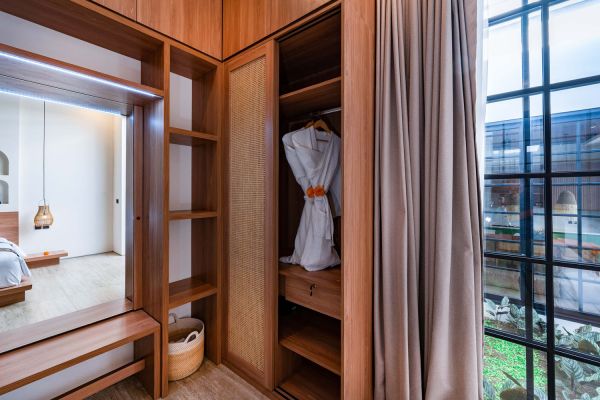 Elegant open wooden wardrobe with bamboo panels in a luxury tropical bedroom, featuring white robes, shelves, a bench, and large window overlooking greenery.